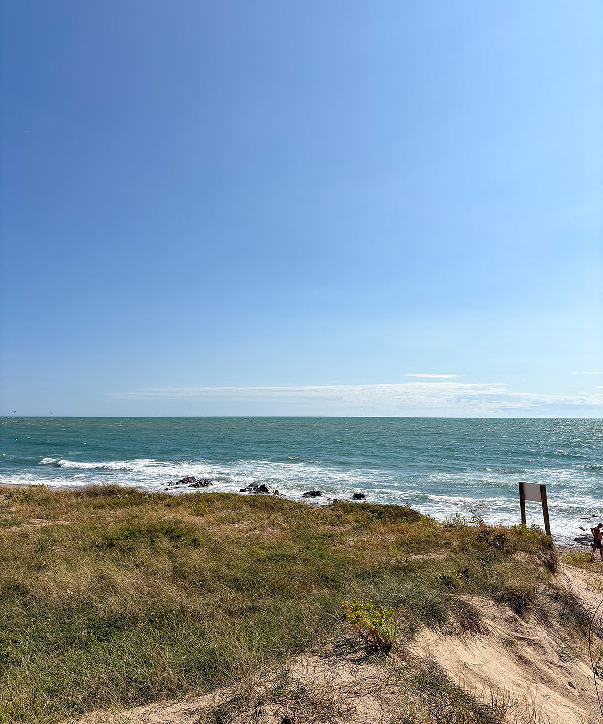 Plage avec dunes et mer agitée sous ciel bleu.